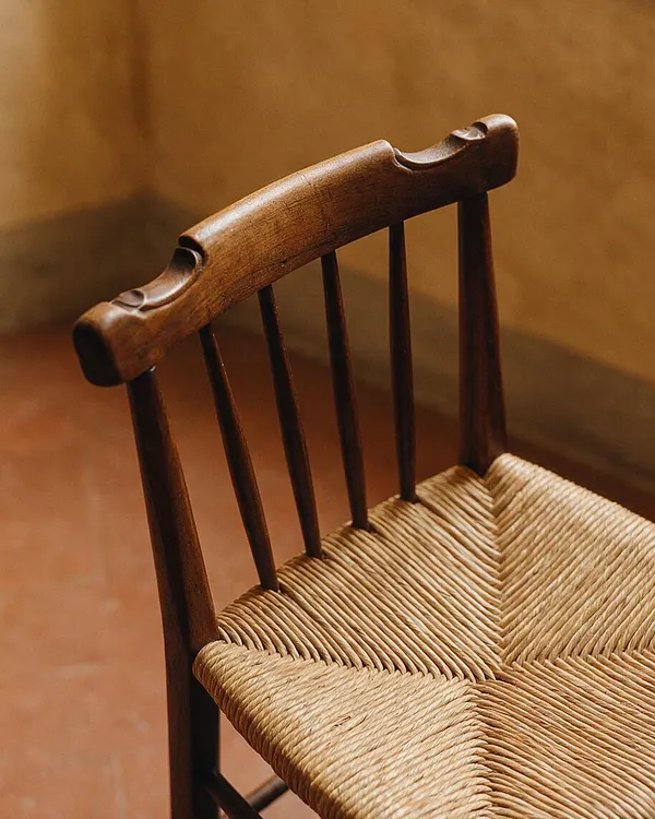 A close-up of a rustic wooden chair with a carved top rail and slender dark spindles. The seat is woven from natural fiber cord in a warm beige tone, set against a softly blurred terracotta and ochre background.