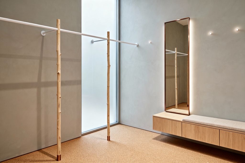 A minimalist home gym with cork flooring, smooth greige plaster walls, and a floor-to-ceiling frosted window. A slim white bar spans the room, fixed to the wall with round brackets and supported by two natural wood poles capped in copper. A floating light-oak bench with a light gray cushion sits below a tall, backlit mirror in a slim copper frame, while two small wall pin lights add soft ambient illumination.