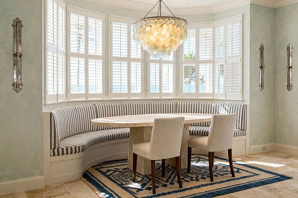 Bright coastal dining nook with a curved built-in banquette upholstered in navy-and-cream stripes surrounding an oval travertine table. White plantation shutters line a bay window with ocean views, a tiered capiz shell chandelier hangs above, two upholstered side chairs rest on a navy geometric flatweave rug, sea-glass green walls hold antiqued mirror sconces, and the floor is tumbled light-beige stone.