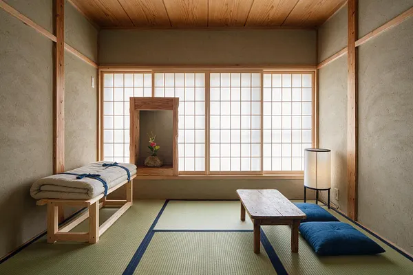 A calm Japanese Zen master bedroom with tatami mat flooring, shoji screens across the back wall, and a low wooden table centered on the floor. A simple bench with folded bedding sits to the left, while blue floor cushions and a paper floor lamp add softness on the right.