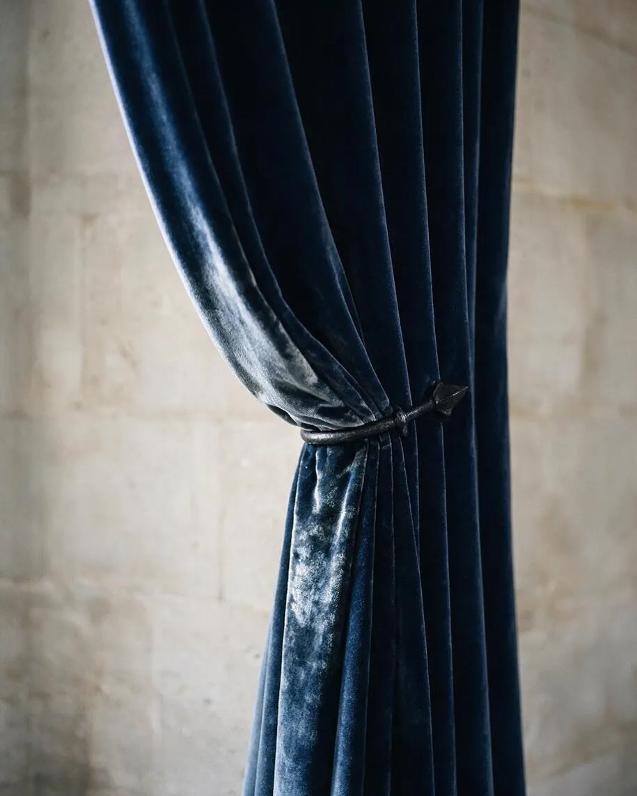 A close-up view of heavy dark blue velvet curtains gathered at the center with a small tieback. The plush fabric catches the light with a soft sheen, set against a muted stone or plaster wall in a moody Gothic-inspired interior.