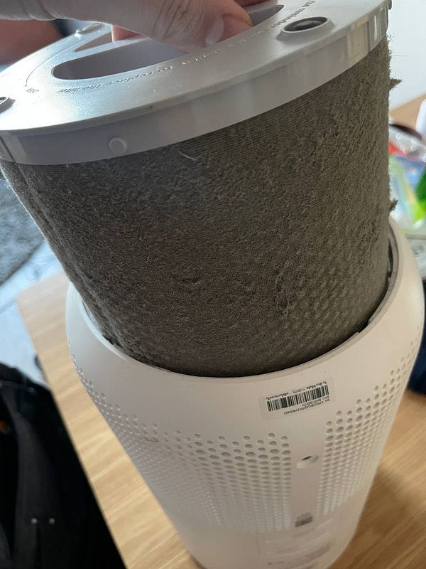A hand is holding a dirty air filter above an air purifier.