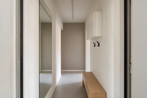 A narrow minimalist entryway with white walls, a large vertical mirror on the left, a light wood bench on the right, and two black wall hooks beneath a white cabinet. A soft taupe accent wall at the end adds depth to the bright, clean hallway.