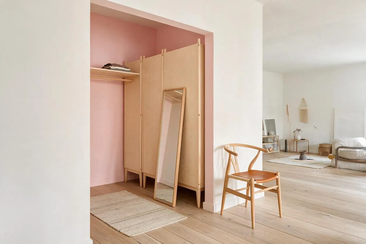 A Scandinavian-style closet nook with pale oak cabinet doors, a slim leaning mirror, and a blush pink back wall. The space opens into a bright minimalist room with light wood floors, a woven rug, and a wooden chair in the foreground.