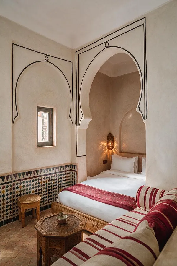 A compact guest bedroom with Moroccan-style arches, cream plaster walls, and a white bed tucked into a recessed alcove. The room includes patterned zellige-style tile wainscoting, a carved wooden side table, a small stool, a red woven bed runner, striped cushions, and a warm lantern wall light.