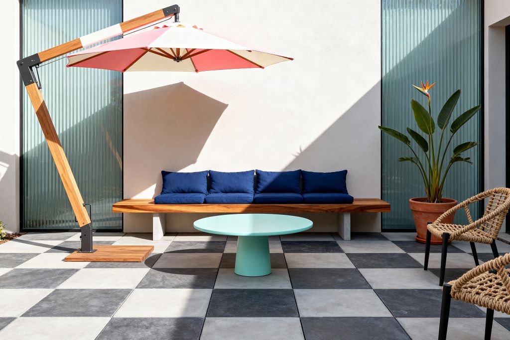 A sunlit patio with large black-and-light-gray checkerboard pavers features a built-in teak bench lined with deep navy seat and back cushions and a mint green round pedestal table beneath a cantilevered umbrella with a wood arm and coral-and-cream canopy. To the right are two woven rope chairs with black metal legs, while ribbed glass panels flank an off-white stucco wall and a terracotta pot holds a bird of paradise plant.