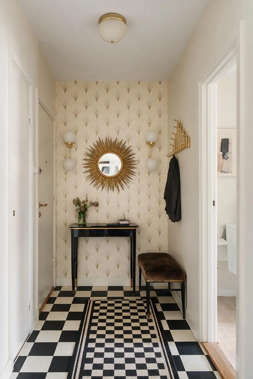 A narrow Art Deco entryway with cream fan-pattern wallpaper, a black-and-white checkerboard floor, and a slim black console table against the far wall. The space features a gold sunburst mirror, globe wall sconces, a brass coat rack, a brown velvet bench, and a checkerboard runner rug.