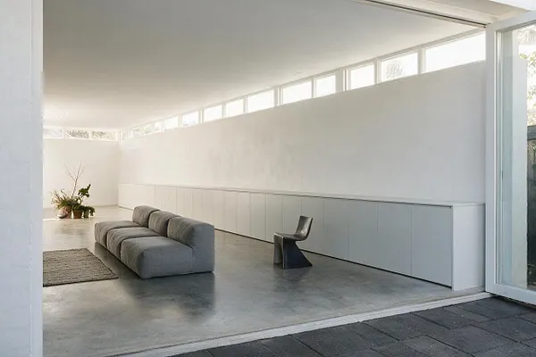 A minimalist backyard lounge with smooth concrete flooring, white built-in storage or bench cabinetry along one wall, a low gray modular sofa, and a small sculptural black chair. Narrow clerestory windows and an open glass door bring in bright natural light, while a small plant arrangement adds a subtle organic accent.