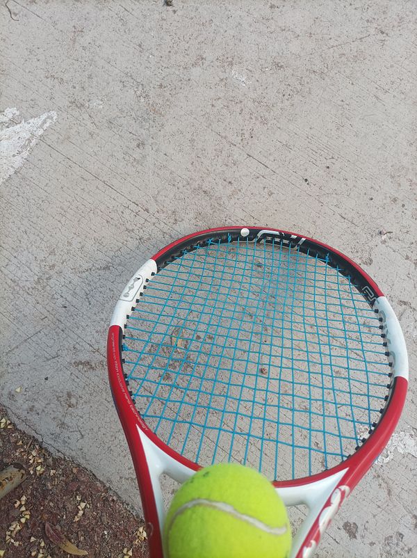 A tennis racket is positioned above a tennis ball on a concrete surface.