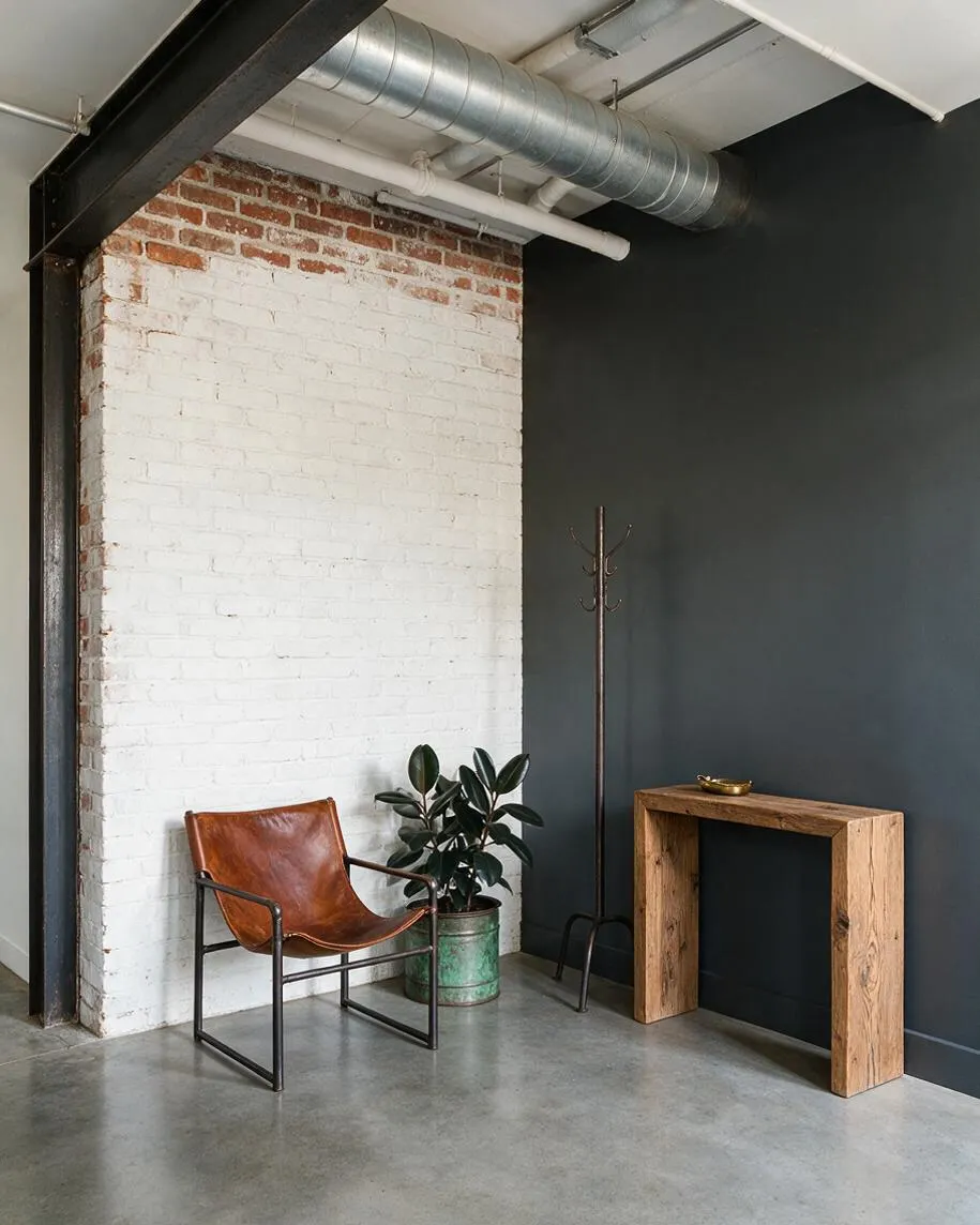 An industrial entryway with a whitewashed exposed brick wall, dark charcoal walls, and visible ceiling ductwork and pipes. A leather sling chair, potted plant, slim metal coat rack, and rustic wood console create a simple, urban welcome area.