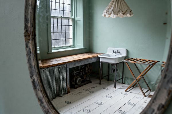A small laundry room, seen through a distressed round mirror, features pale sage walls and trim, a leaded-glass window, and soft daylight. A reclaimed wood counter with a green-and-white gingham skirt runs beneath the window, next to a white wall-mounted porcelain sink on metal pipe legs. A folding wooden drying rack stands to the right, while white painted floorboards with gray-blue stencils and a pleated fabric pendant create a gently worn, Shabby Chic atmosphere.