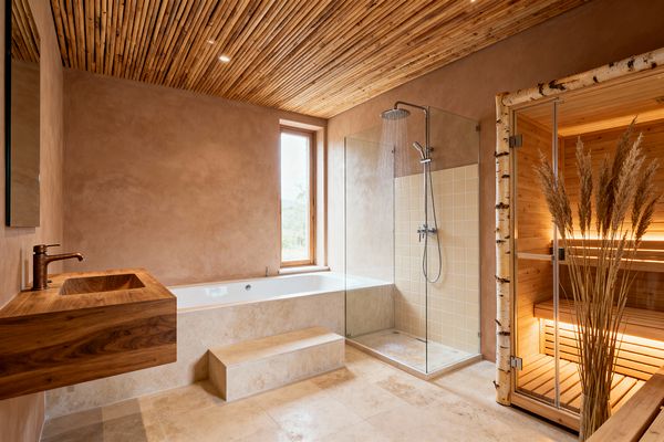 A Scandinavian bathroom with warm clay-plaster walls, a slatted timber ceiling with recessed lights, and pale travertine flooring. On the left, a floating solid-wood vanity with a copper faucet sits beside a built-in white bathtub and stone step. A glass walk-in shower with cream square tiles and chrome fixtures adjoins a birch-wood sauna with soft backlighting and a glass door, while a tall window brings in daylight.