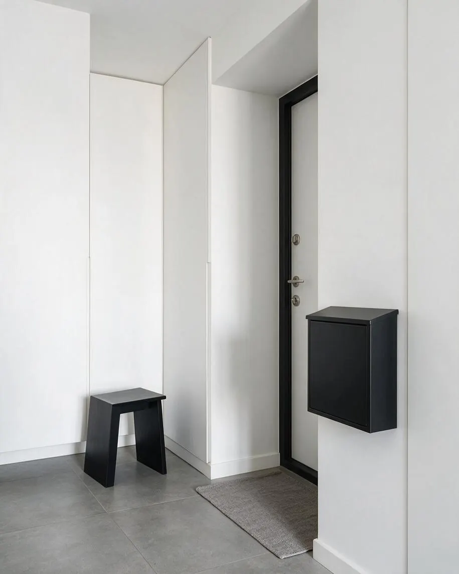 A minimalist front porch with smooth white walls, large gray floor tiles, and a black-framed front door. A small black stool sits in the corner, and a sleek black wall-mounted mailbox and gray doormat add simple, functional details.