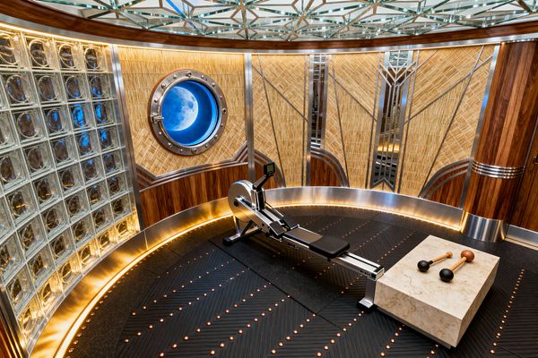 A curved Art Deco home gym centers a sleek rowing machine on black rubber flooring with chevron grooves and dotted accent lights. Left is a wall of glass blocks; ahead, straw marquetry panels with chrome geometric inlays and rich walnut trim curve around the room. A polished metal porthole glows blue with a moon image. A mirrored, faceted ceiling and perimeter LED cove lighting add shine. A light travertine plinth holds two vintage-style Indian clubs.