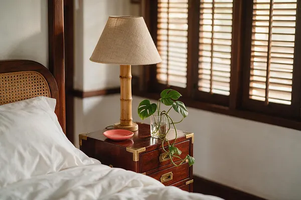 A tropical-style bedroom vignette with a bamboo table lamp on a dark wood bedside cabinet. The cabinet has brass corner accents and drawers, and sits beside a bed with white bedding and a cane headboard, with warm sunlight filtering through wooden shutters.