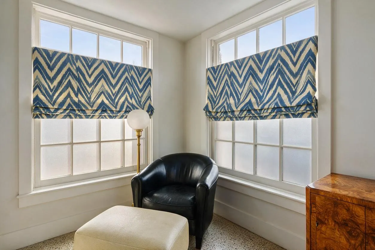 A bright sunroom corner with two large windows fitted with blue-and-cream chevron Roman shades. The space includes a black leather club chair, a cream ottoman, a brass floor lamp with a globe shade, and a burled wood cabinet, all set against light walls and trim.