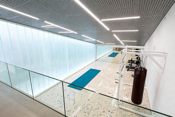 A long, minimalist home gym features a glowing backlit wall of vertical channel glass, a perforated gray ceiling with linear LED strips, and a speckled terrazzo floor. Teal exercise mats lie along the corridor, while a powder-coated white steel rig holds a brown leather punching bag beside a mirrored wall with weight bench and plates; a clear glass balustrade borders the upper level, creating an open, airy feel.