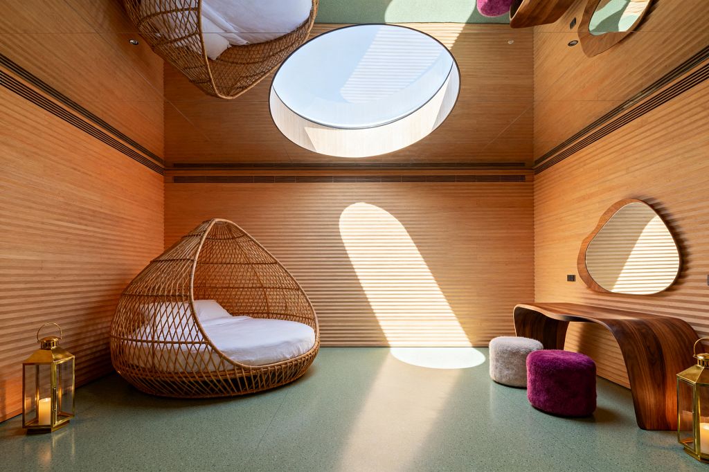 A children’s room with ribbed honey-toned wood walls and a green speckled floor features a large circular skylight that casts an elongated pool of sunlight. A rattan cocoon bed with white bedding sits near a sculpted walnut desk and an amoeba-shaped mirror, with magenta and cream plush poufs and brass lanterns; a mirrored ceiling reflects the furnishings and skylight, heightening the playful, surreal atmosphere.