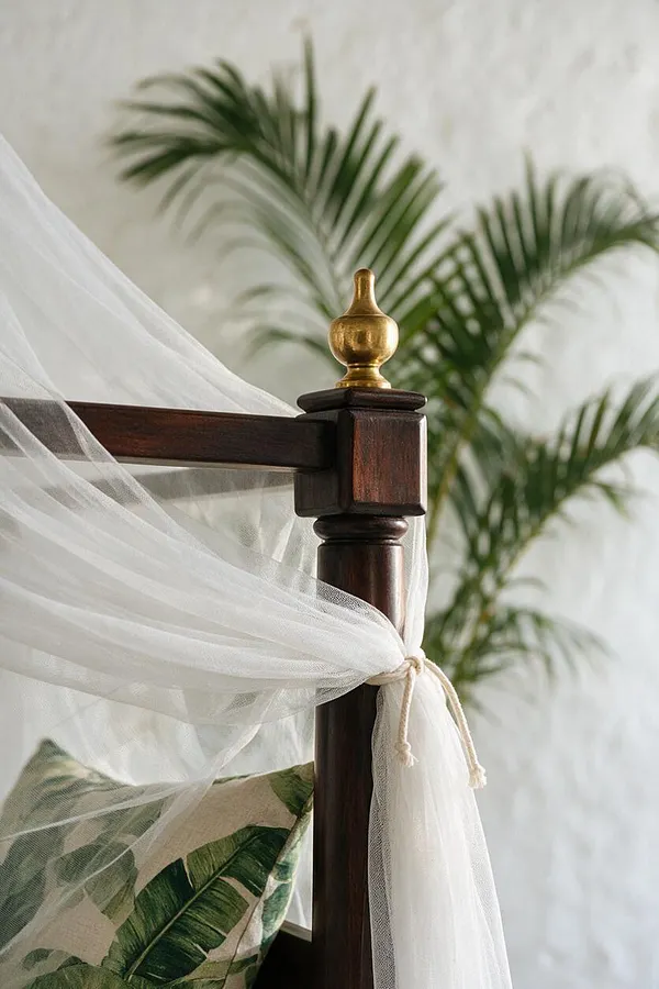 A close-up of a dark wooden bed post topped with a brass finial, with sheer white canopy fabric tied softly around it. In the background, blurred palm leaves and a tropical leaf-print pillow add a breezy, resort-like feel.
