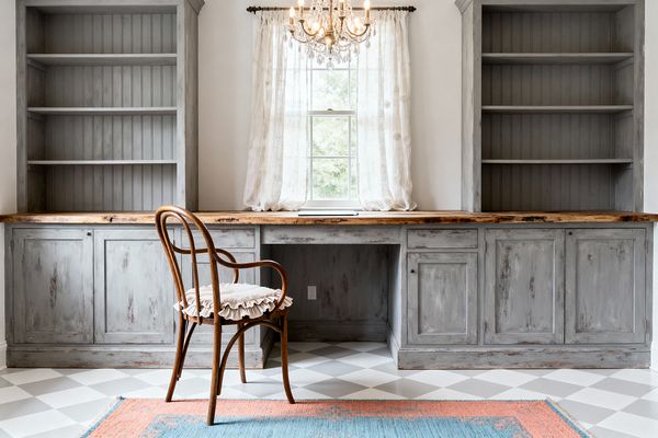 Symmetrical home office with distressed gray built-in cabinets and beadboard shelving flanking a central window dressed in sheer, subtly patterned curtains. A long natural wood desktop spans the wall, lit by a crystal chandelier, with a curved bentwood chair and ruffled cushion in front; a light gray and white checkerboard floor and a coral-blue flatweave rug add pattern and color in a bright, vintage-inspired space.