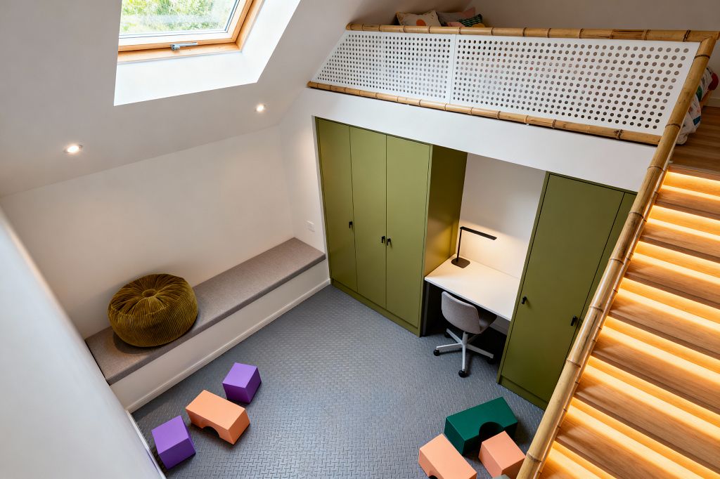 A compact, lofted kids’ room with a sloped ceiling and wood-framed skylight features olive-green built-in wardrobes framing a white desk with a gray swivel chair and black task lamp, all set on a gray textured rubber floor scattered with colorful foam blocks. A warm LED-lit oak stair with bamboo trim leads to a loft behind a perforated white guard, while a long bench with a gray cushion and mustard pouf sits below recessed spotlights.