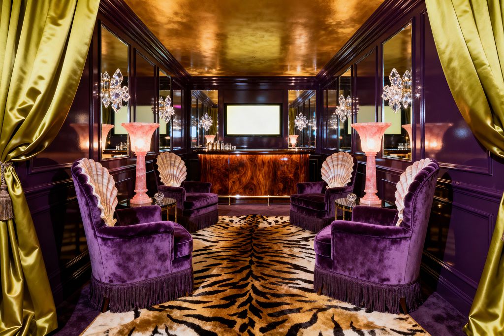 A glamorous media room with glossy aubergine paneled walls and mirrored inserts features four purple velvet fringe armchairs with scallop-shell backs arranged symmetrically on a plush tiger-stripe rug. Pink alabaster torchiere lamps, crystal starburst sconces, and an underlit burl-wood bar glow beneath a reflective gold ceiling, while chartreuse silk curtains frame the entrance and a wall-mounted screen anchors the far wall.