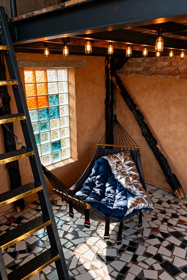 An intimate, rugged bedroom with a suspended hammock-style bed holding a quilted blue sleeping bag with a reflective silver liner, hung from black straps under a steel mezzanine. Warm Edison bulbs line the underside of the platform, illuminating rough clay-plaster walls, exposed brick, and charred timber braces. A patchwork floor of broken ceramic tiles and a glass-block window with clear, amber, and teal squares complete the post-apocalyptic look.