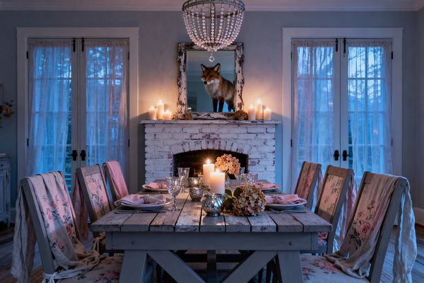 A candlelit shabby chic dining room with a weathered wood trestle table set for six, blush napkins, vintage glassware, and dried hydrangeas. Distressed floral-upholstered chairs surround the table; behind, a whitewashed brick fireplace holds pillar candles beneath a distressed mirror reflecting a fox figure, while a crystal-bead chandelier hangs above and sheer lace curtains soften the French doors against blue twilight.