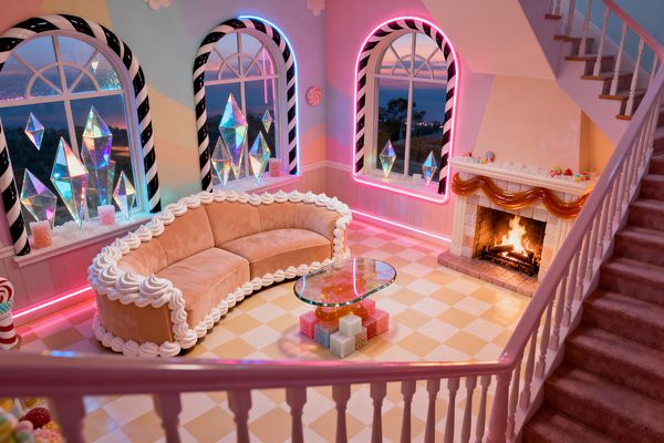 A whimsical living room in pastel tones features a curved peach velvet sofa trimmed with whipped-cream piping, a glass coffee table set on glittery cube bases, and a butter-and-cream checkerboard floor. Arched windows with black-and-white licorice frames glow with pink and aqua LED neon, displaying iridescent crystal sculptures. A stone-tile fireplace burns beneath a glossy caramel-like garland, while a pink balustrade and mauve carpeted stair rise at right.