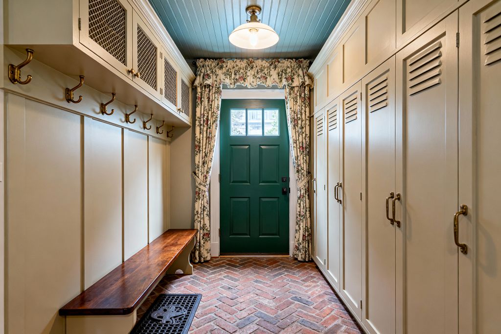 A narrow traditional mudroom with creamy built-in cabinetry: louvered lockers with brass pulls on the right, and a dark-stained wood bench with brass hooks and mesh-front upper cabinets on the left. The red herringbone brick floor leads to a deep green paneled door framed by cream floral curtains, while a muted blue beadboard ceiling and a schoolhouse pendant light create a warm, classic atmosphere.