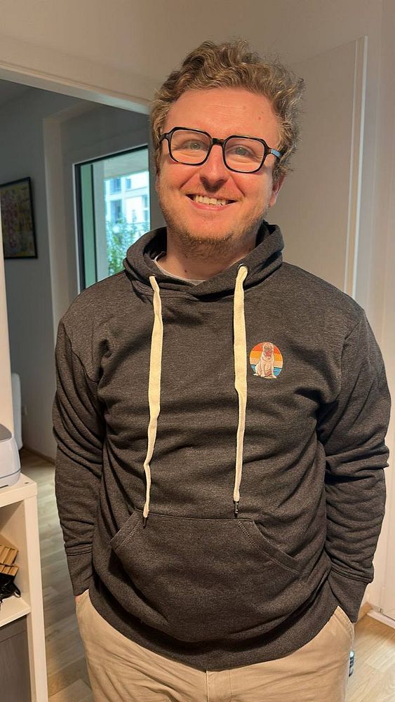 A smiling man wearing a dark hoodie with a colorful logo stands in a well-lit room.