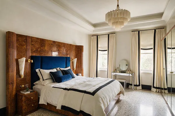 A master bedroom with an Art Deco-inspired look featuring a large burl wood headboard wall, a deep blue velvet headboard, and a white bed dressed with black trim bedding and layered pillows. Cream curtains, mirrored closet doors, a glass vanity, and a tiered chandelier add elegance, while terrazzo flooring and brass accents complete the room.