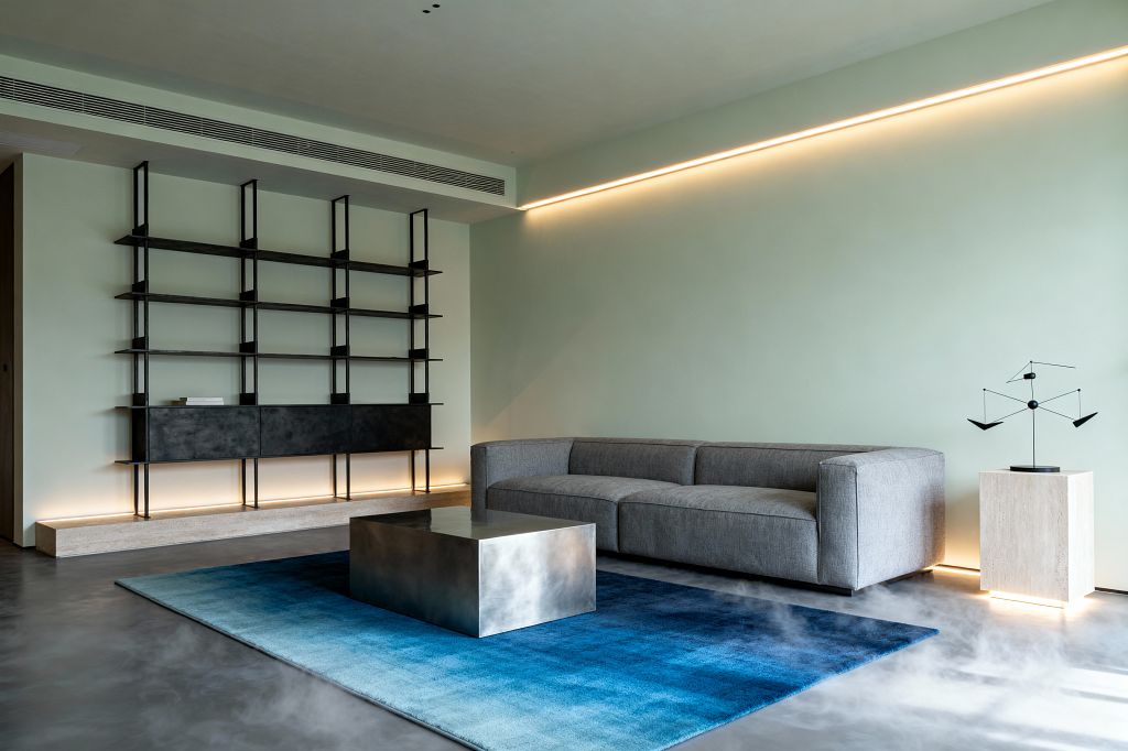 A minimalist living room features a pale sage wall washed by a long linear LED. A low gray sectional is paired with a reflective rectangular steel coffee table on a blue gradient rug over smooth gray microcement flooring. At the side stands a light travertine pedestal supporting a small black mobile. Opposite, black powder-coated steel shelving with a closed lower cabinet appears to float above a travertine plinth, accented by base lighting and a slim ceiling air vent.
