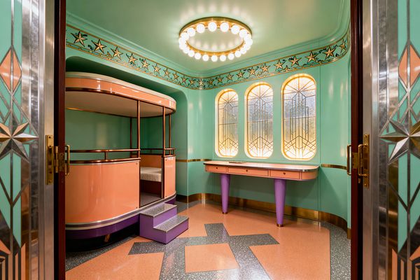 A kids’ room in Art Deco style features mint walls with a star frieze and a circular brass ceiling light ringed with globe bulbs. A curved peach bunk bed with brass rails and lilac steps faces three arched stained-glass windows backlit with warm light, beside a peach console on purple tapered legs. The terrazzo-pattern floor in salmon and charcoal and geometric metal inlays on the doors complete the period look.