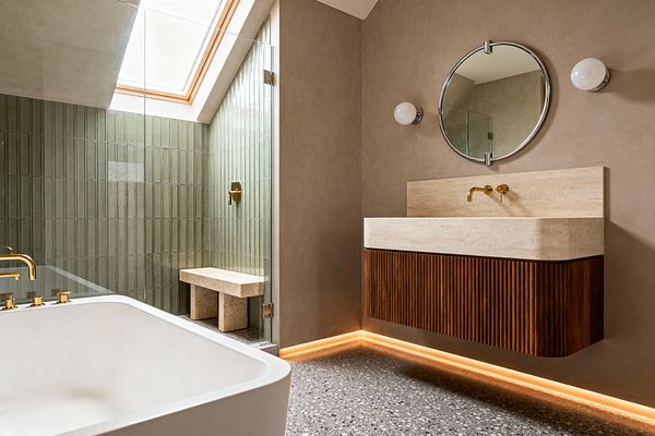 A transitional bathroom features a floating fluted-wood vanity topped with a thick travertine basin and a wall-mounted brass faucet beneath a round mirror and globe sconces. At left, a glass-enclosed shower lined with vertical sage-green tiles includes a travertine bench and brass fittings under a skylight. A white soaking tub with brass taps faces terrazzo floors, while warm LED toe-kick lighting washes taupe plaster walls with a soft glow.