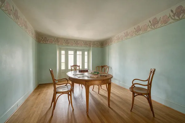 A sparse game room with pale mint walls, a floral Art Nouveau border near the ceiling, and a polished round wooden table surrounded by bentwood-style chairs. Light wood floors and a bright windowed door create a soft, airy atmosphere.