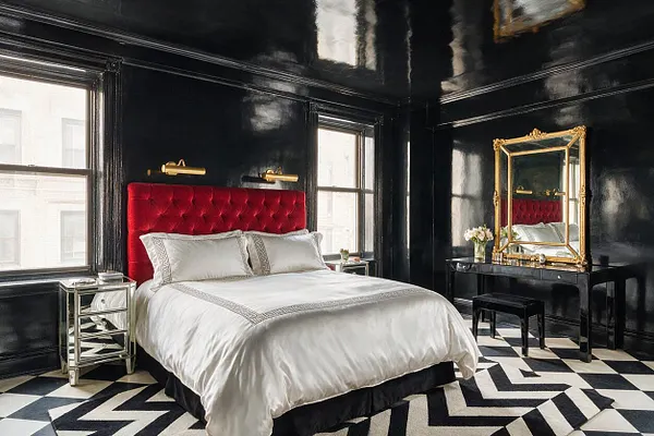 A dramatic master bedroom with glossy black walls and ceiling, a red tufted velvet headboard, and white bedding with Greek key trim. Mirrored nightstands, a black vanity with a gold-framed mirror, and a black-and-white geometric rug complete the Hollywood Regency look.