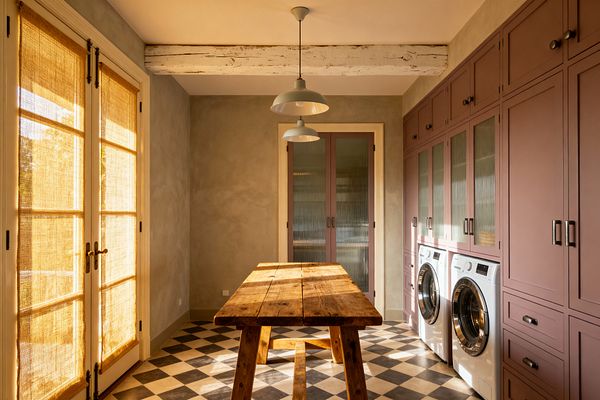 A narrow laundry room features dusty mauve built-in cabinets along one wall, with reeded glass doors and a white front-loading washer and dryer set. A reclaimed wood work table sits on black-and-cream checkerboard tile, while limewashed gray walls, a distressed white ceiling beam, sage-green metal pendants, and French doors with woven natural shades fill the space with warm afternoon light.