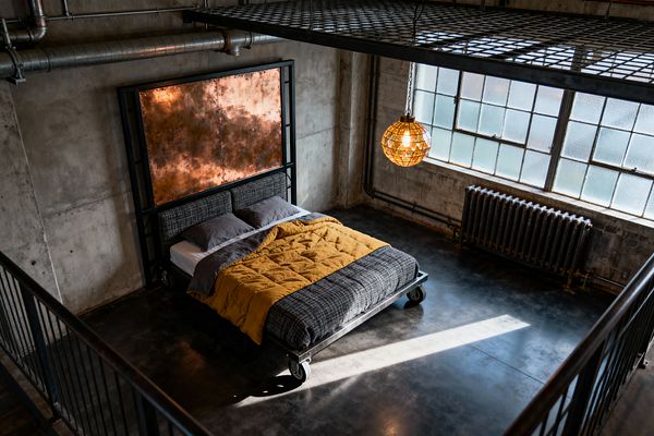 An industrial loft bedroom features a black steel bed on large casters with gray tweed upholstery, set against a tall riveted copper panel headboard. Sunlight pours through steel-framed factory windows with textured glass, illuminating polished concrete floors, exposed ductwork, visible conduits, and a cast-iron radiator. A spherical cage pendant glows amber beside raw concrete walls, with a steel-grate mezzanine overhead and charcoal and mustard bedding adding warmth.