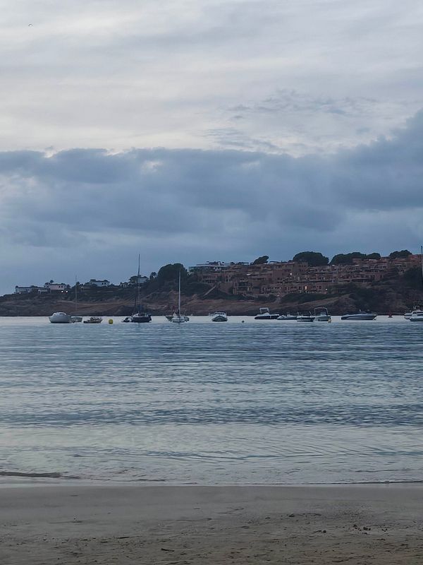 A calm sea view with several boats anchored near the shoreline and a residential area in the background under a cloudy sky.