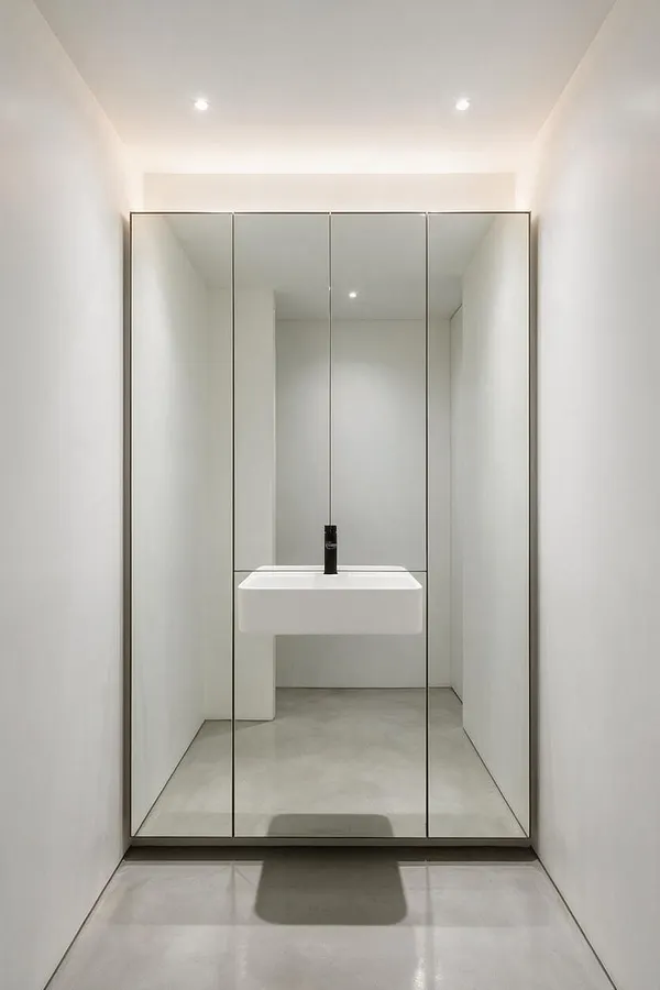 A narrow minimalist powder room with white walls, a floating white sink, and a large mirrored cabinet spanning the back wall. Thin dark metal framing divides the mirror panels, while recessed ceiling lights and a pale polished floor keep the space bright and spare.