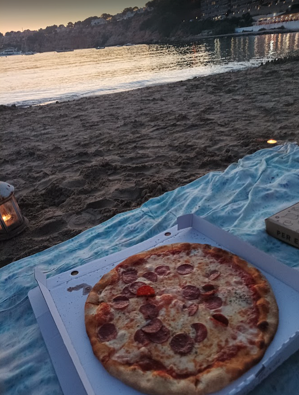 A pizza is placed on a sandy beach with a scenic view of the water at sunset.