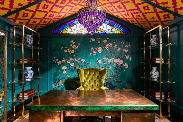 A richly colored home office features an oxidized copper desk facing a tufted chartreuse velvet chair, set against a teal wall with a cherry-blossom chinoiserie mural. Brass-framed glass shelves with blue-and-white porcelain and leather-bound books flank the scene, while a magenta and yellow patterned ceiling and an amethyst crystal chandelier glow beneath a multicolored stained-glass transom, creating a layered, maximalist look.