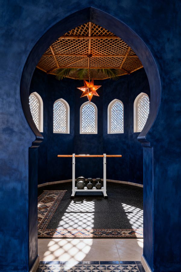 A view through a keyhole-shaped arch shows a compact home gym with deep cobalt plaster walls and an octagonal layout. Five arched windows with white lattice screens cast patterned light onto black speckled rubber flooring edged by a patterned rug. A wooden ballet-style barre on a white metal stand faces a row of kettlebells. Above, a woven rattan ceiling with palm fronds centers a glowing brass-and-amber Moroccan star pendant.