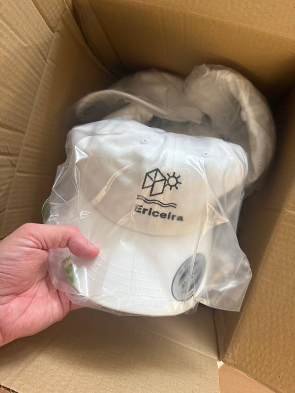 A hand is holding a white cap with an 'Ericeira' logo inside a cardboard box.