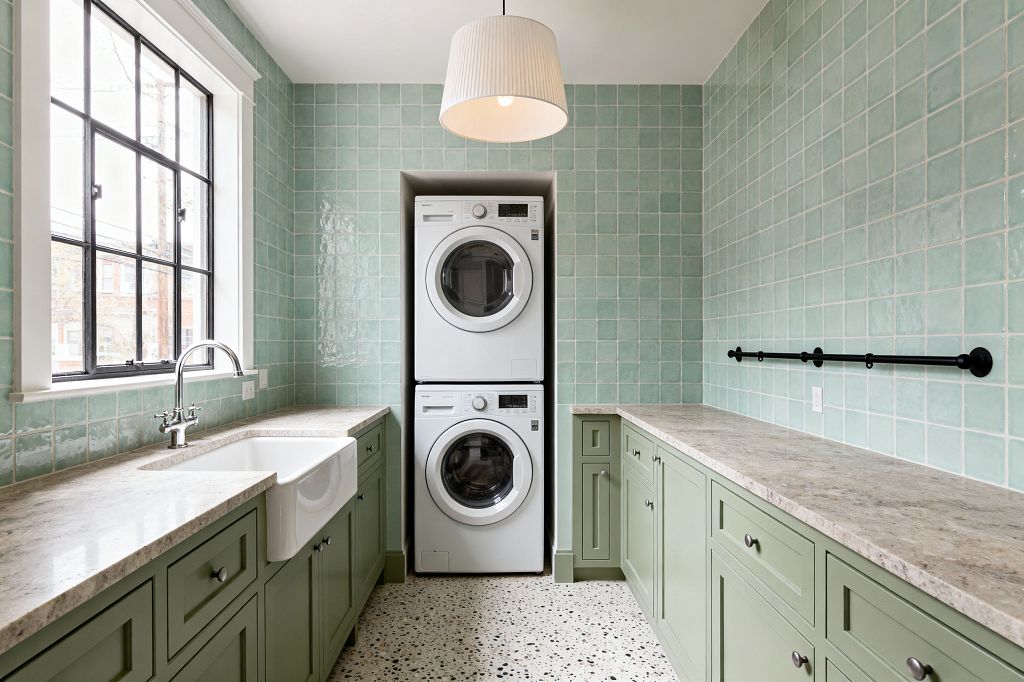 A compact galley laundry room with a stacked white washer and dryer centered in a niche, flanked by sage green Shaker cabinets and light stone countertops. A white apron-front sink sits beneath a black-grid window. Glossy mint square tiles clad the walls to the ceiling, and terrazzo flooring with black flecks runs underfoot. A matte black wall-mounted drying rod lines the right wall, and a white drum pendant provides overhead light.