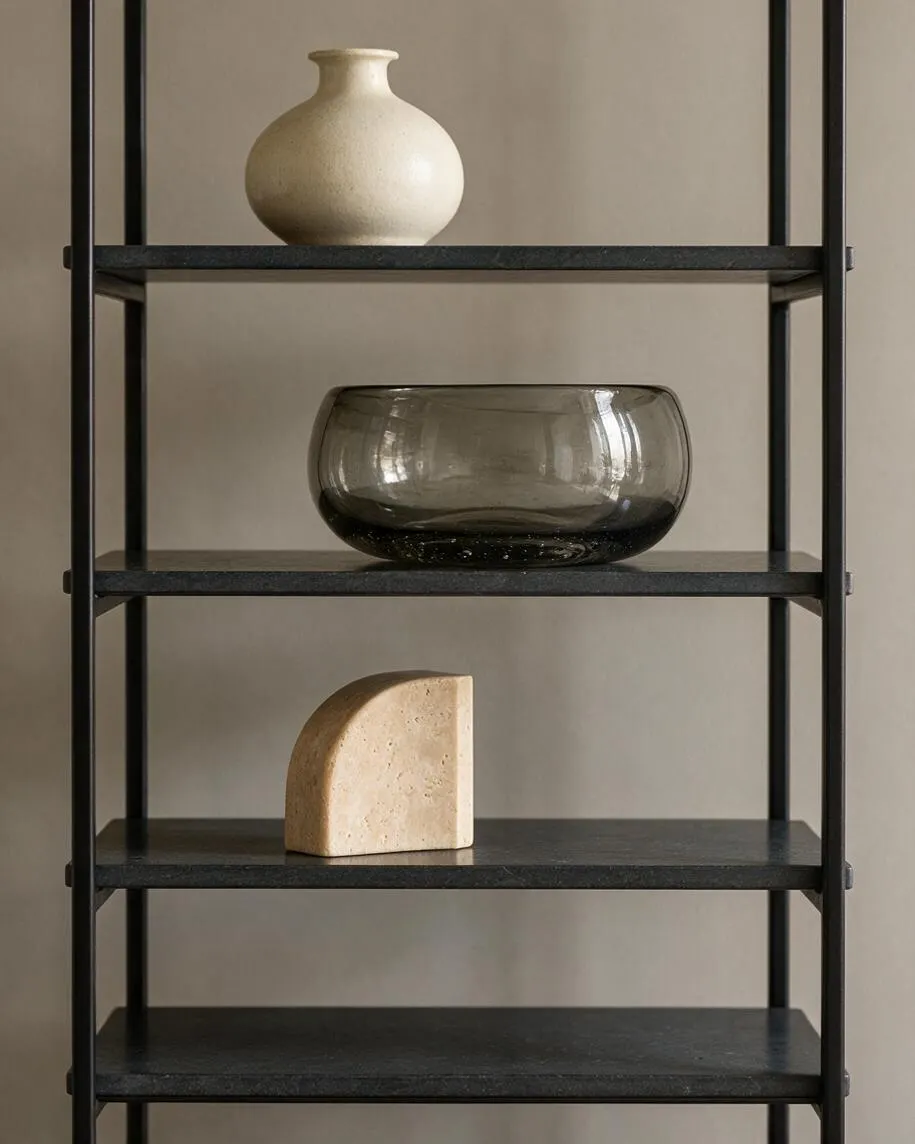 A modern black metal shelving unit against a warm beige wall holds three sculptural decorative objects: a cream ceramic vase on the top shelf, a smoked glass bowl on the middle shelf, and a small stone-like abstract form on a lower shelf. The composition is minimal and balanced with soft natural light.
