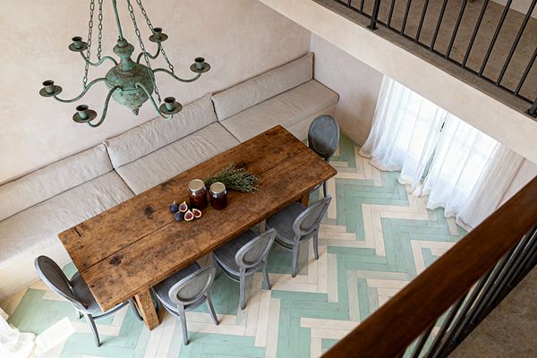 Overhead view of a French Country dining room with a long reclaimed-wood table flanked by weathered gray oval-back chairs and a built-in linen banquette along a plaster wall. A verdigris wrought-iron chandelier hangs above, while a painted herringbone floor in mint green and creamy white, sheer white curtains, and a black iron railing create an airy, rustic setting; jars of preserves, figs, and herbs style the tabletop.