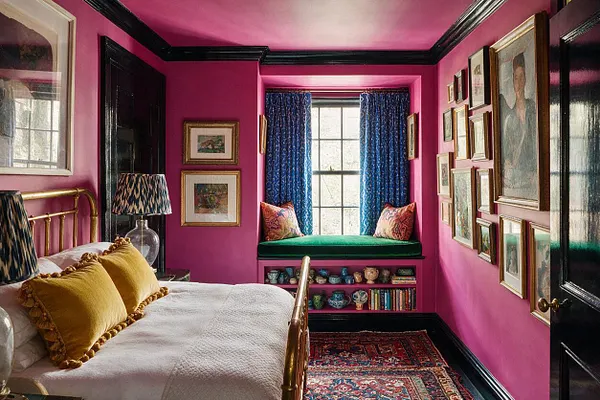 A vibrant guest bedroom with hot pink walls, black trim, and a brass bed dressed in white bedding and mustard pillows. The room features a window seat with blue curtains, a gallery wall of framed art, patterned lampshades, layered rugs, and shelves filled with decorative objects.