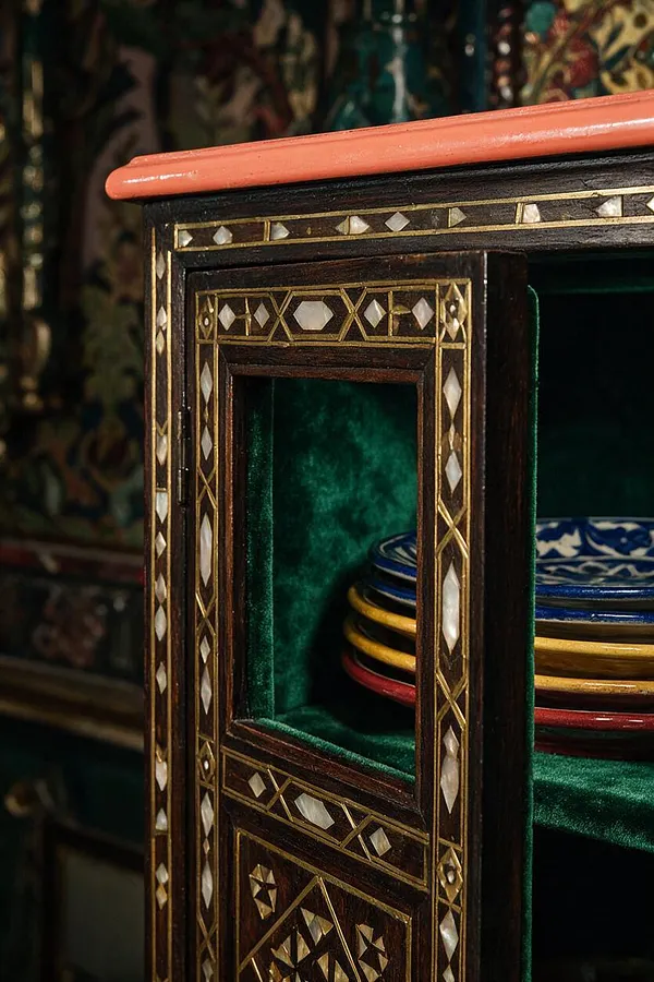 A close-up of an ornate dark wood cabinet with intricate geometric inlay in gold and mother-of-pearl. The cabinet has a coral-colored top edge, a rich emerald velvet interior, and stacked blue, yellow, red, and white patterned plates on one shelf.